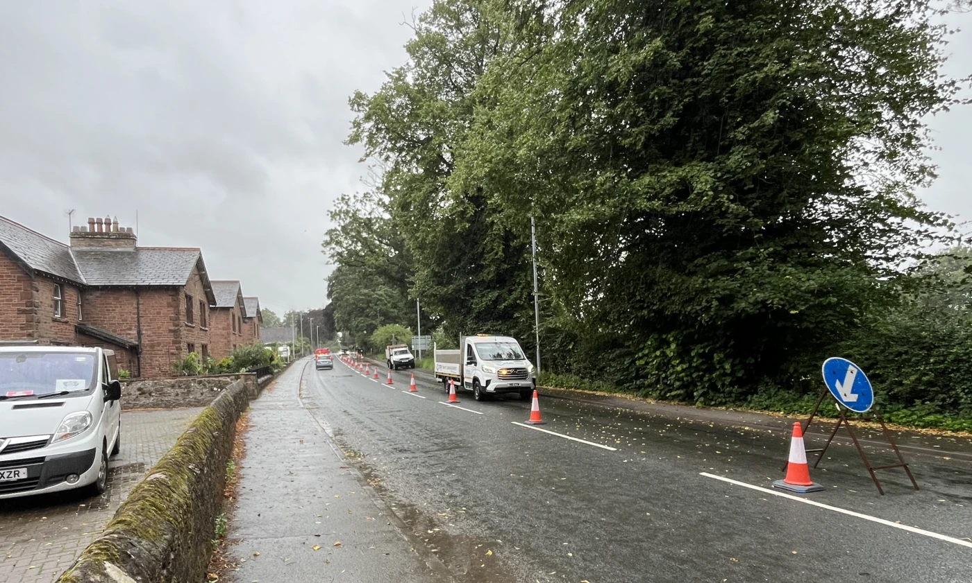 Coned lane closure on a wet road beside a line of mature roadside trees, with a tipper truck standing in the closed lane